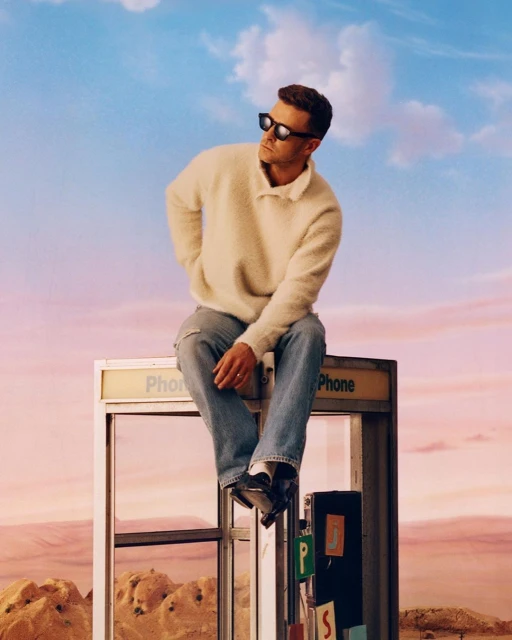 Adult male in sunglasses sits atop a vintage phone booth, wearing a cream sweater and jeans against a desert and sky backdrop.