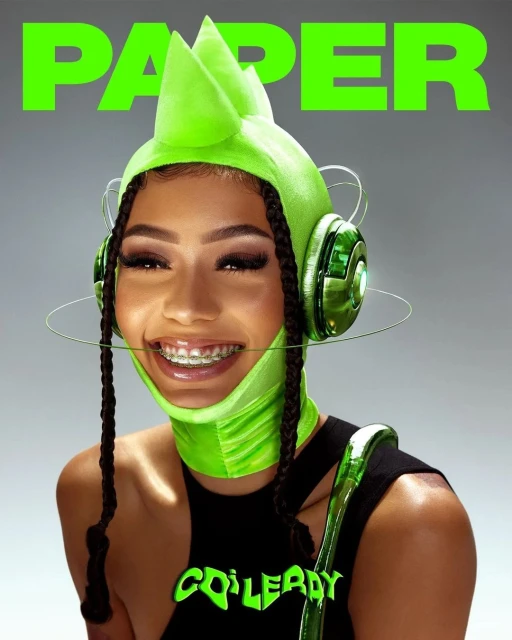 Close-up shot of a smiling woman with braids wearing braces, a neon green hat-mask, and a black top, featuring the word 'PAPER' on top.