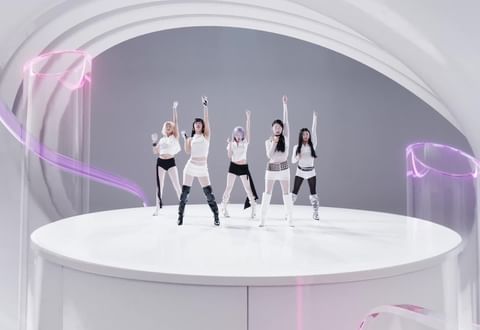 Five women in coordinated white outfits pose on a white platform under studio lighting with purple translucent design elements, smiling.