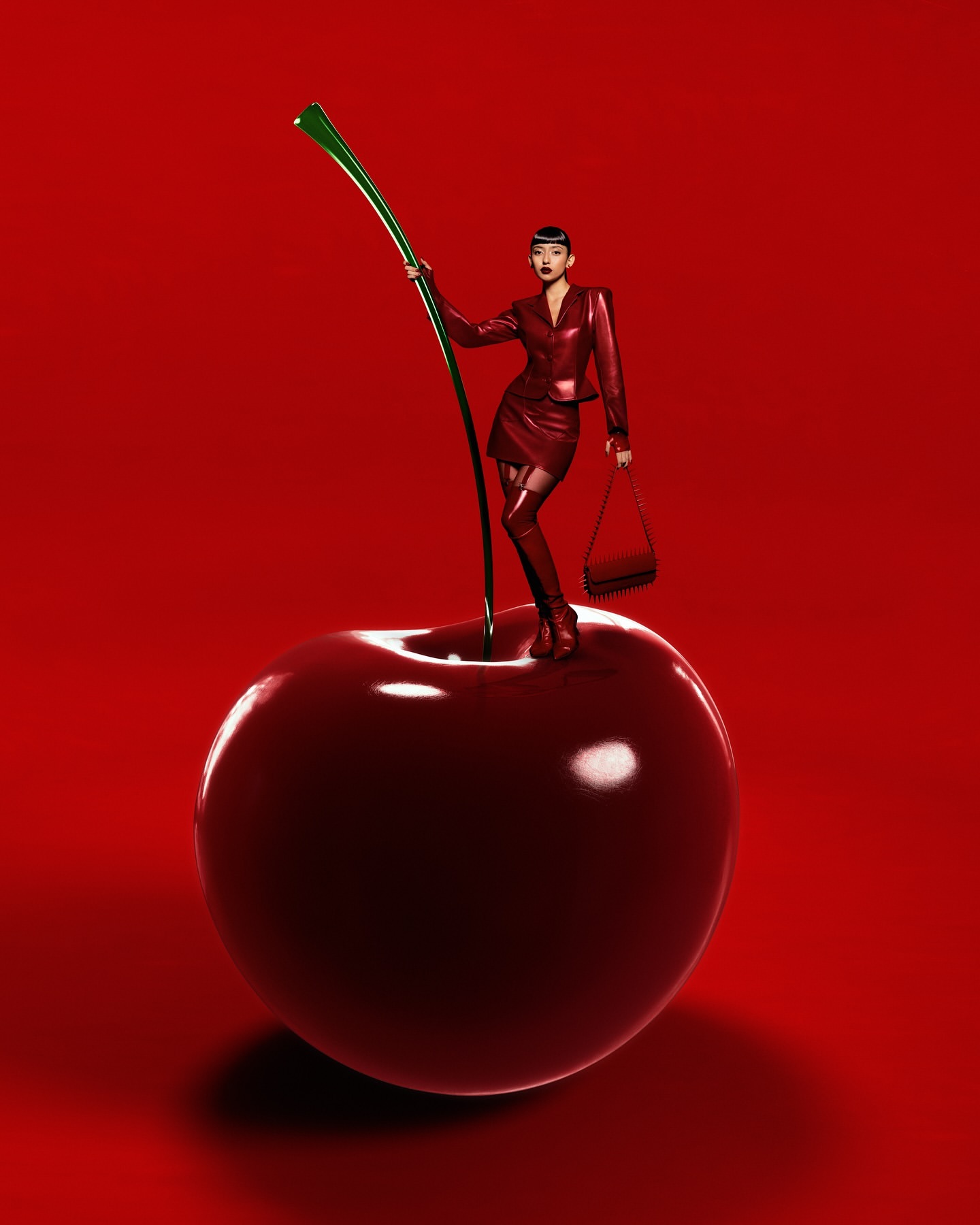 Adult woman in red skirt suit standing on a large cherry with a green stem against a red background in a studio shot.