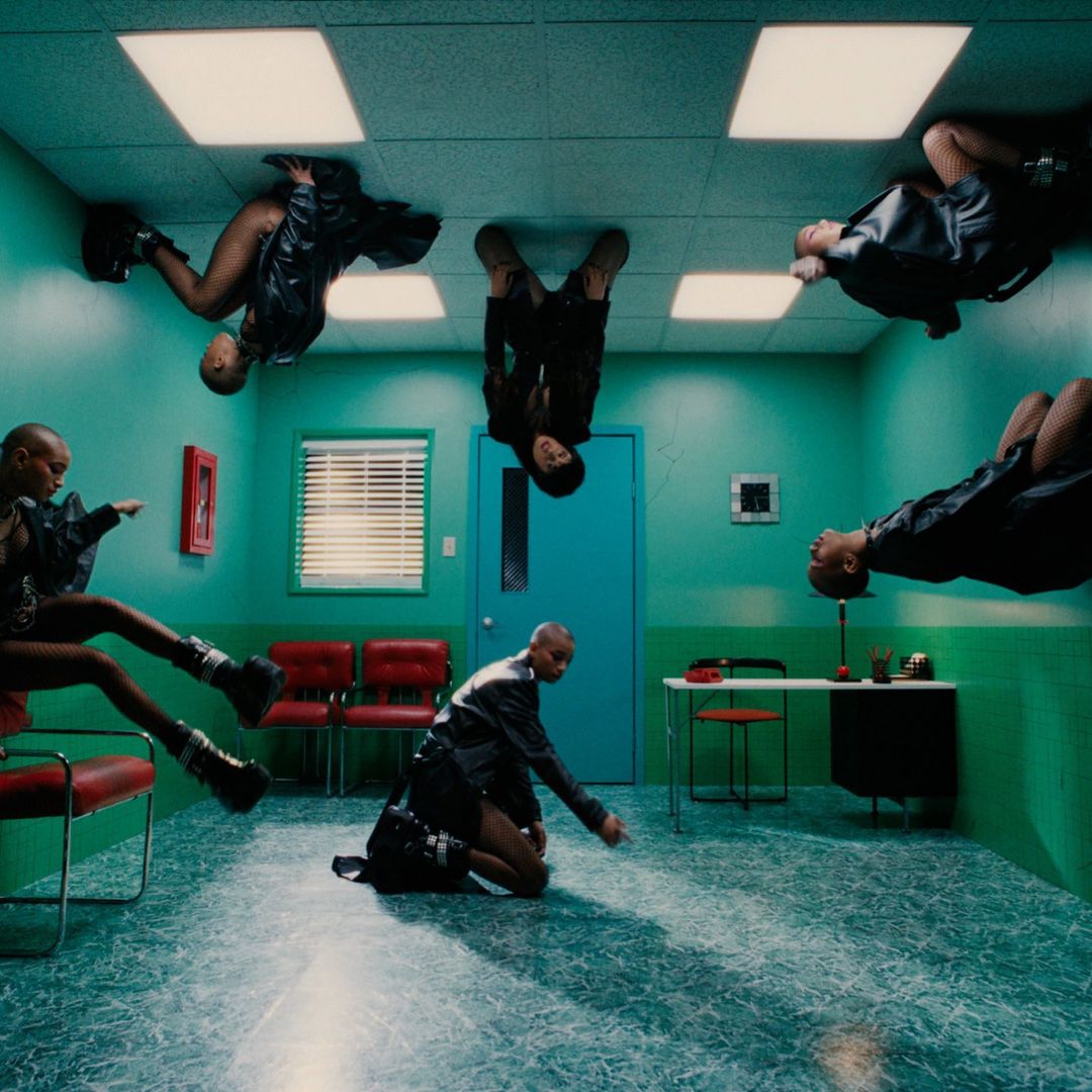 Five bald people in a room: one kneeling, the others seemingly on the ceiling, wearing black leather jackets and boots in a bizarre pose.