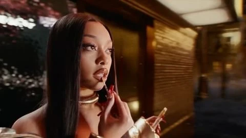 Glamorous woman applies lip gloss indoors, wearing a gold necklace, long red nails, and an off-the-shoulder gold dress in soft lighting.