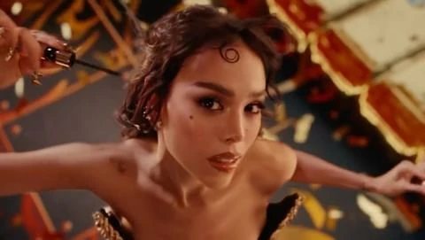 Close-up of a model applying mascara; head-and-shoulders view with decorative patterned backdrop in neutral and gold tones.