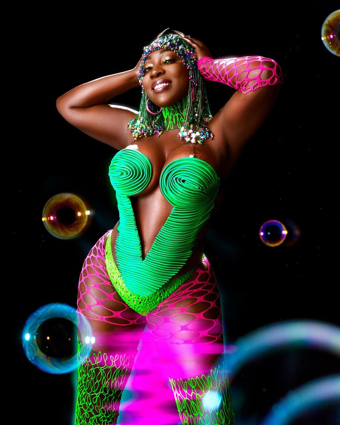 A Black woman with dark skin poses with her hands up wearing a bright green top, pink bottom, and matching beaded head piece on black backdrop.