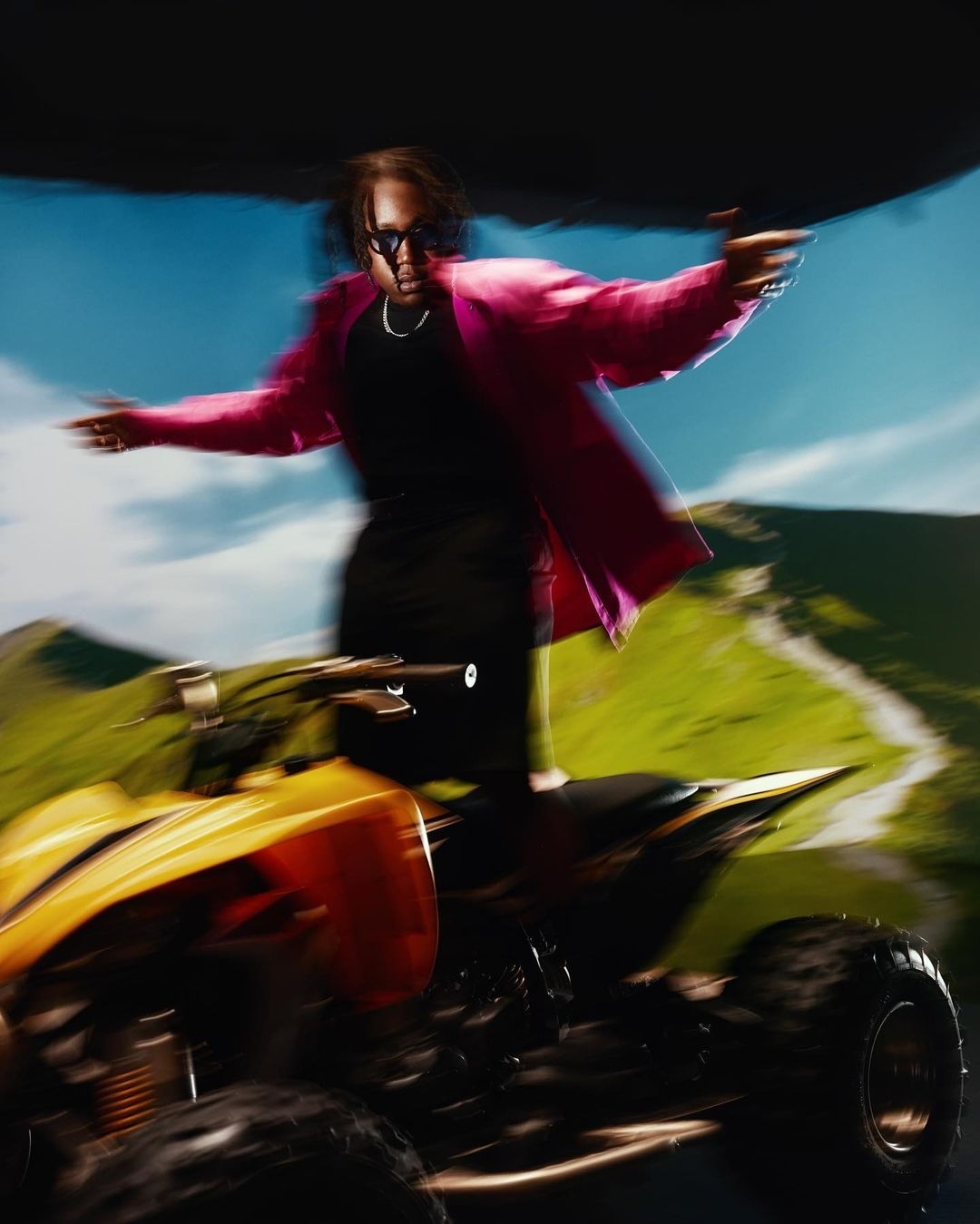 A man in a pink jacket and sunglasses rides an all-terrain vehicle with a blurred background of mountains and sky, creating motion.