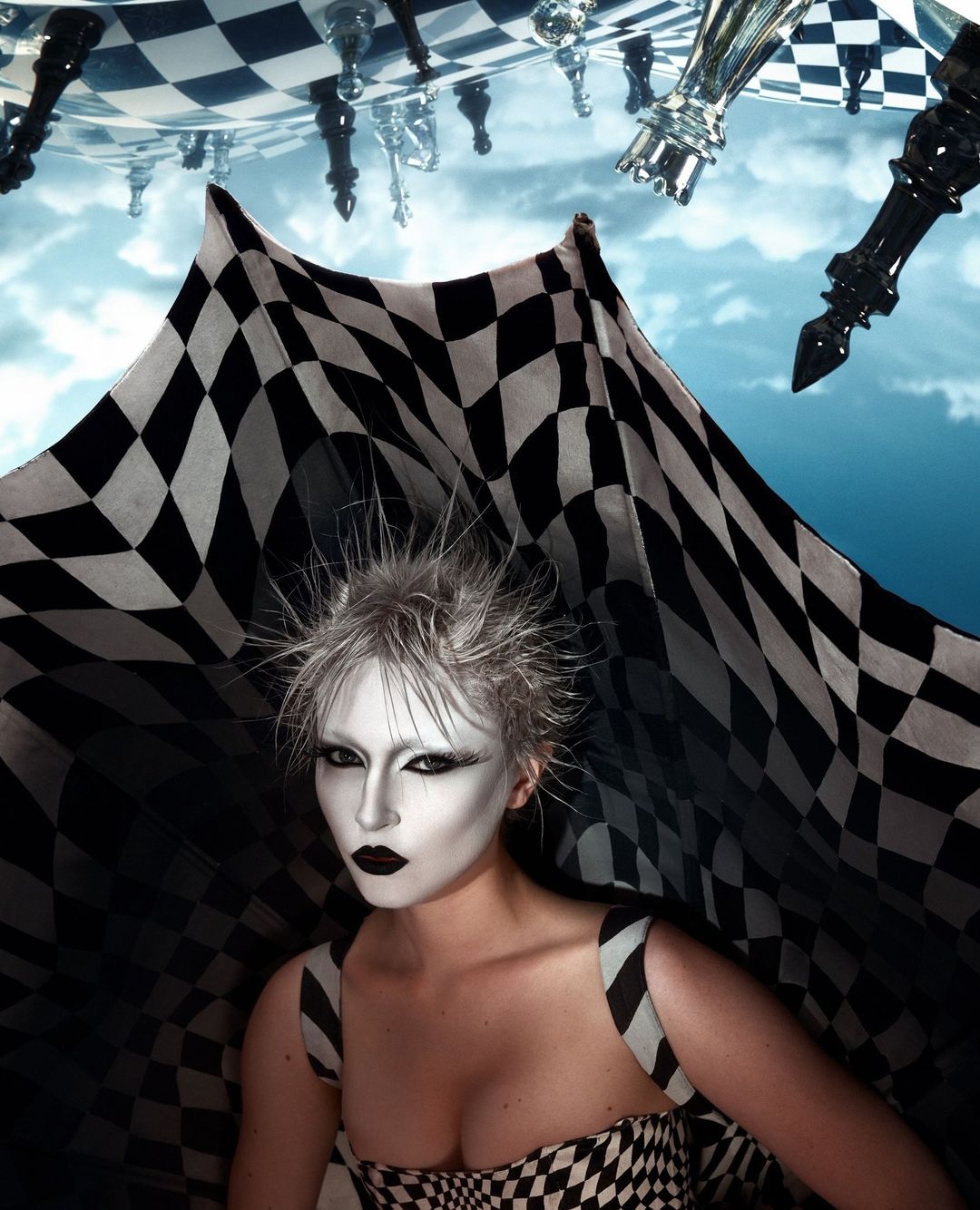 Woman with dramatic makeup poses under floating chess pieces and a checkered cape in an eye-catching beauty and fashion image.