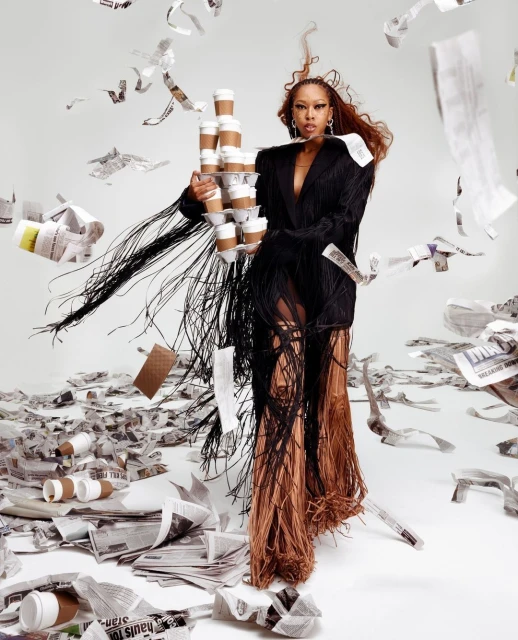 Stylish woman in fringed jacket holding stacked coffee cups amidst scattered newspapers, set against a clean, bright white backdrop.