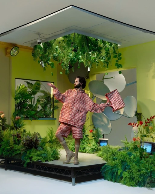 Adult male in patterned shirt and shorts on a stage surrounded by green foliage and screens in a green room under hanging plants.