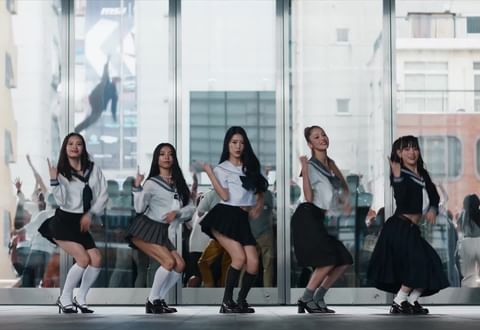 Five Asian women in matching school uniforms perform choreography in front of a glass building with a crowd of people in the background.