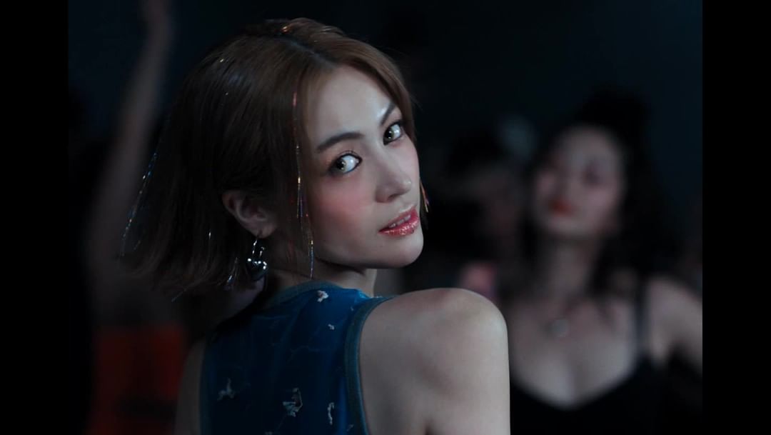 Woman with brown hair, wearing a blue top with confetti-like strands in her hair and dangling earrings, poses in front of a crowd.