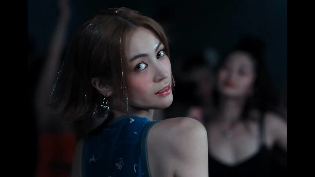 Woman with brown hair, wearing a blue top with confetti-like strands in her hair and dangling earrings, poses in front of a crowd.