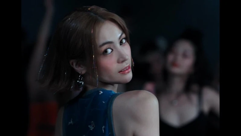 Woman with brown hair, wearing a blue top with confetti-like strands in her hair and dangling earrings, poses in front of a crowd.