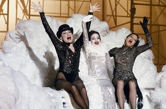 Three adult women perform on stage in matching sequin costumes, black hosiery, and hats. The trio are smiling with raised arms on a fluffy cloud.