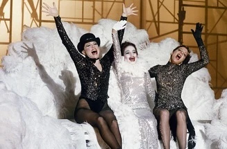 Three adult women perform on stage in matching sequin costumes, black hosiery, and hats. The trio are smiling with raised arms on a fluffy cloud.