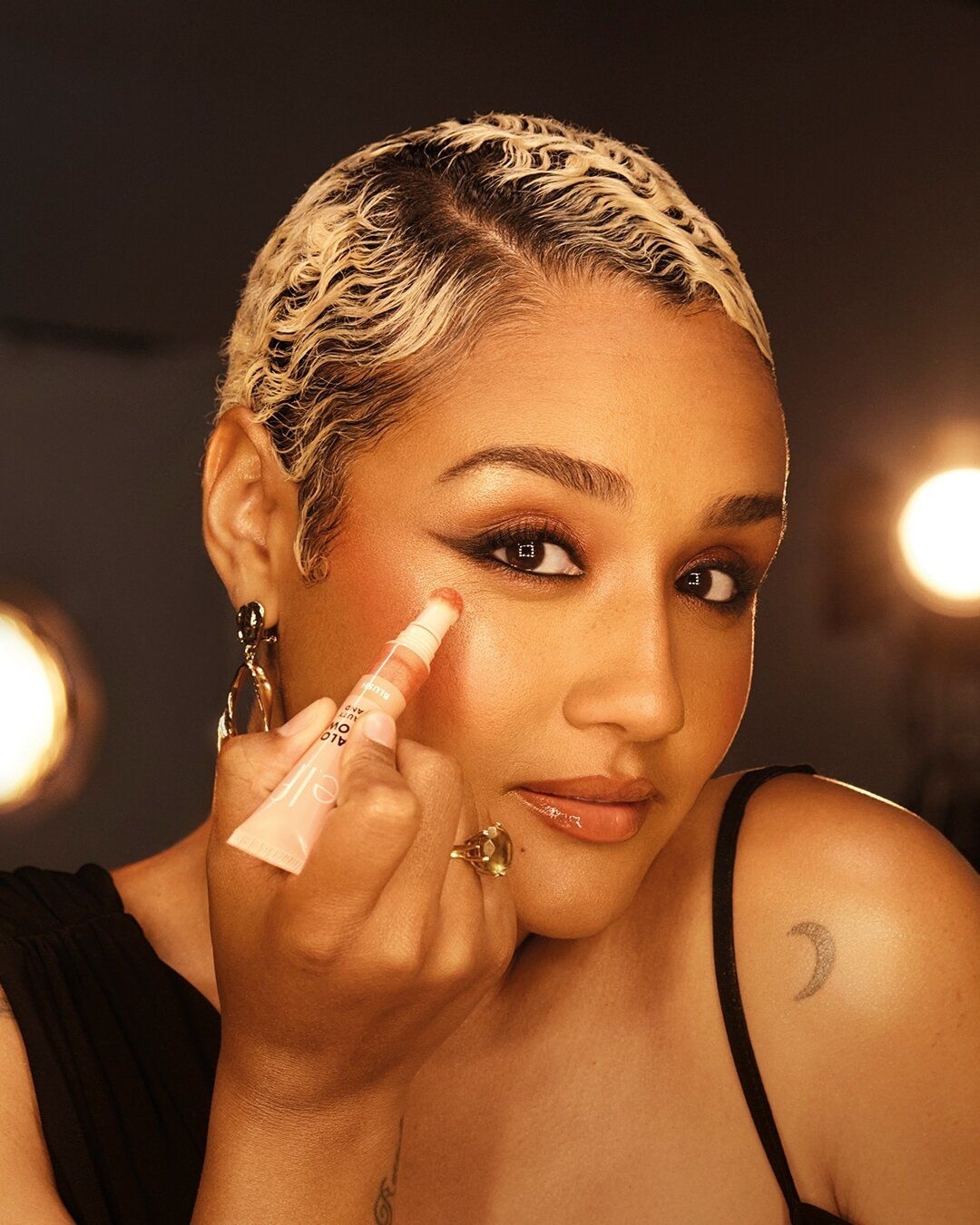 A woman with wavy blonde hair applies makeup to her face in a close-up, beauty shot with warm lighting and a dark background.