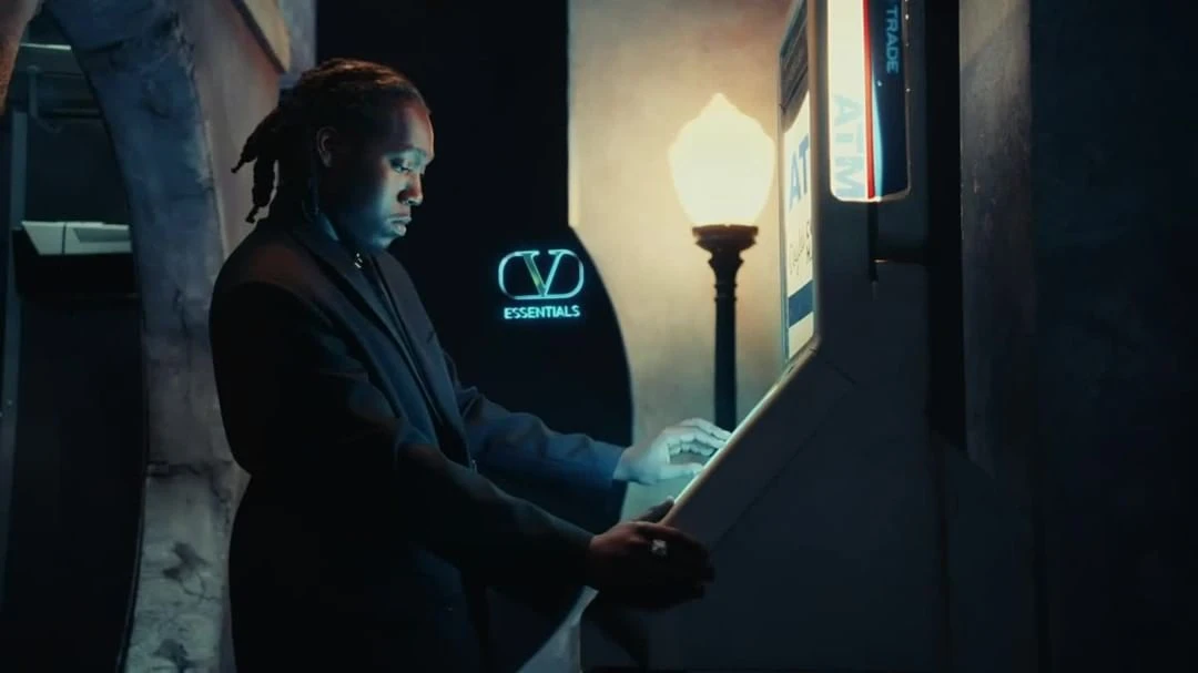 Man in suit interacting with ATM in dim indoor setting; illuminated by neon sign and nearby street light.