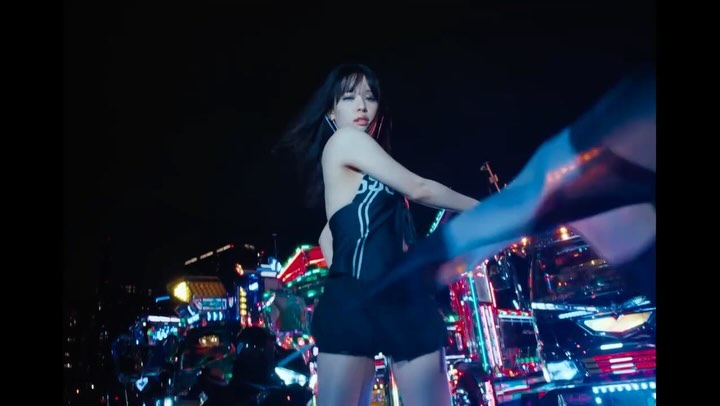 A woman with dark hair poses outdoors at night in a dark halter top and shorts in front of a colorful, reflective cyberpunk vehicle.