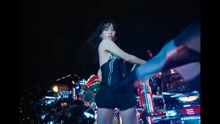 A woman with dark hair poses outdoors at night in a dark halter top and shorts in front of a colorful, reflective cyberpunk vehicle.