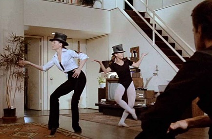 Two women in dance attire and top hats perform a synchronized routine in an indoor living space. Third figure watches from the foreground.