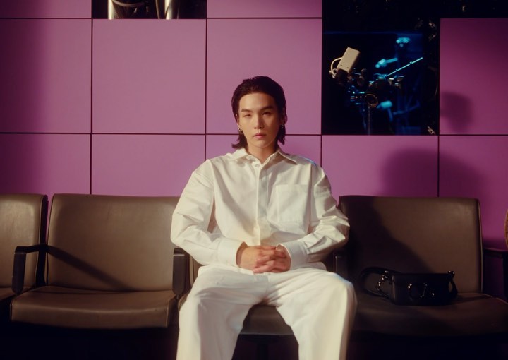 Adult male in a button-down shirt poses while seated on a chair in front of a purple wall, with a professional camera in the background.