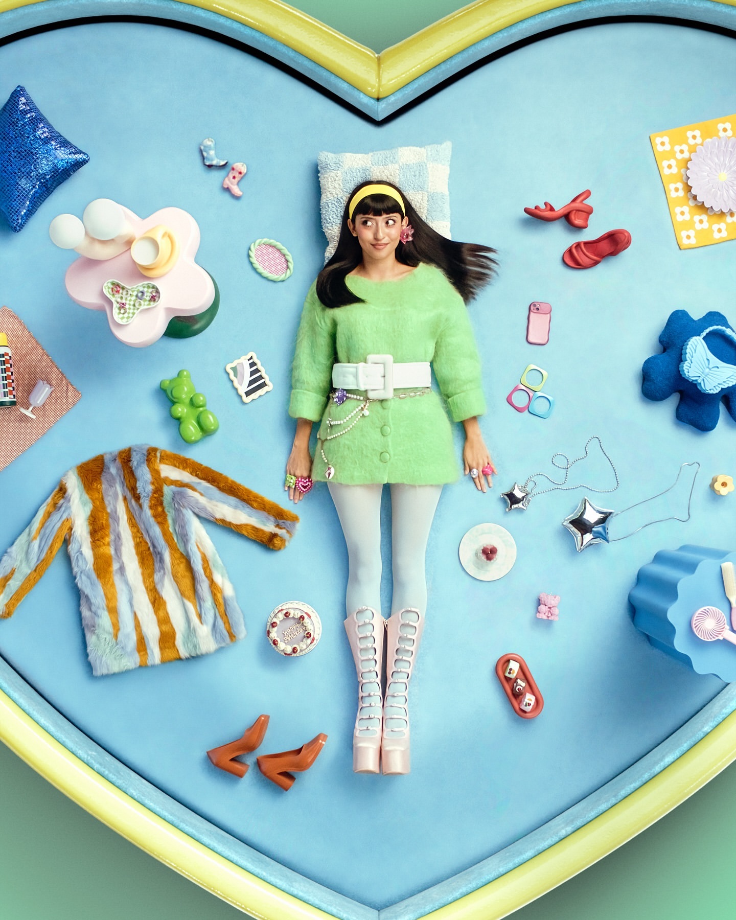 Overhead studio shot of a woman lying on a light blue surface surrounded by toys and props in a whimsical, colorful setting.