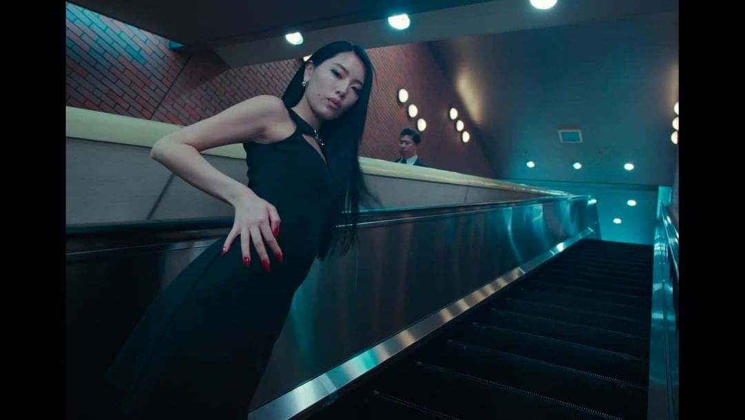 An Asian woman poses in a black dress and red nails next to a silver escalator, showcasing fashion in an urban setting with brick elements.