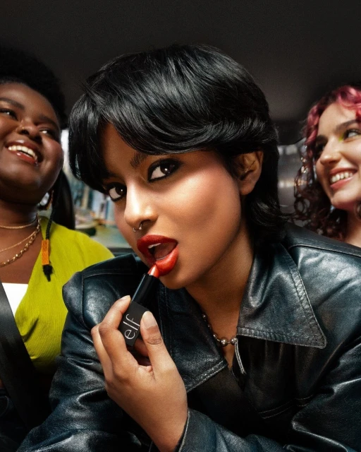 Three women in a car apply makeup. A woman in a leather jacket holds lipstick, others smile in the background with natural makeup.