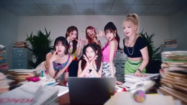 Group of six smiling adult women posing together indoors behind a desk with a laptop, with stacks of files and papers visible.