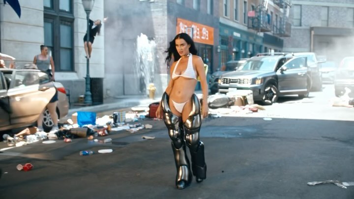 A female model in a white bikini top and futuristic leg coverings walks down a city street strewn with trash and debris.