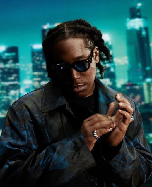 An African-American male wearing sunglasses and a patterned overshirt poses in front of a city skyline backdrop in a portrait-style shot.