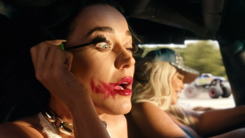 Close-up of a woman applying mascara in a car, with a blonde woman and a monster truck visible in the background.