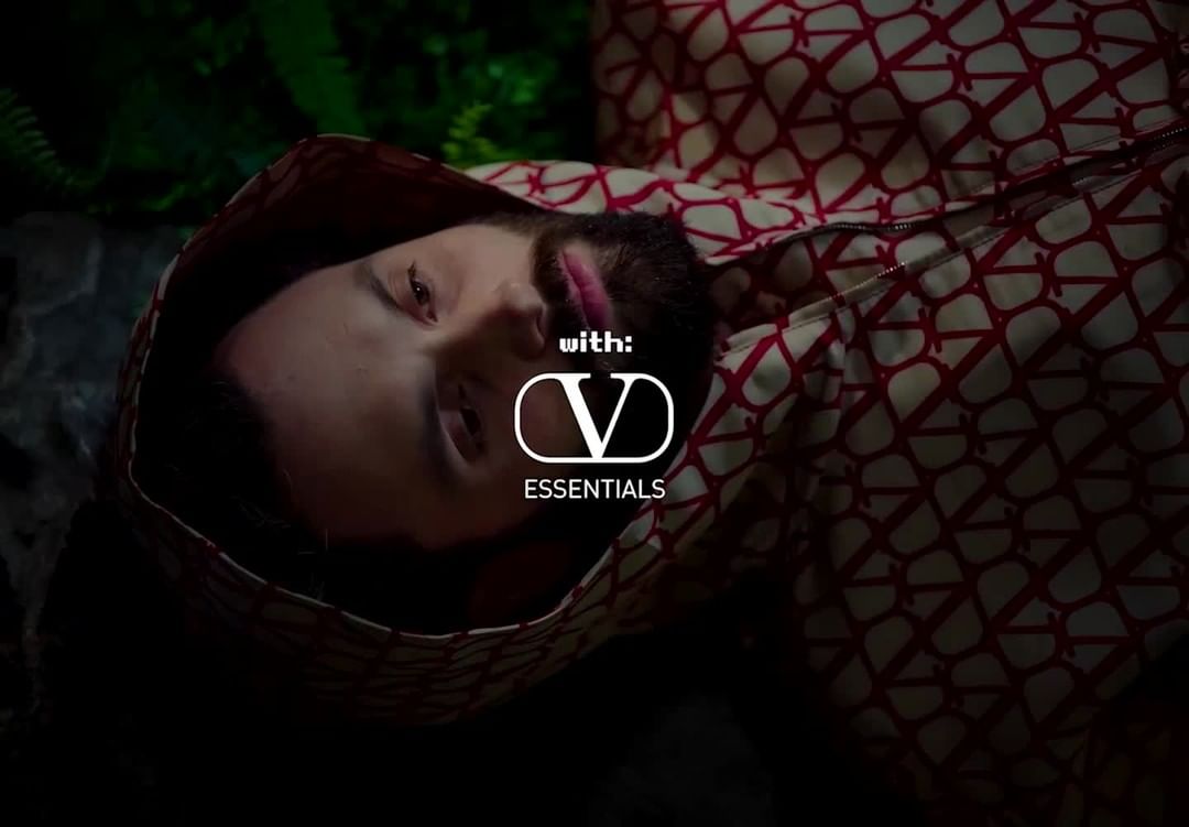 Man with beard and hooded jacket poses against a dark background with green leaves, wearing a printed jacket featuring Valentino logo.