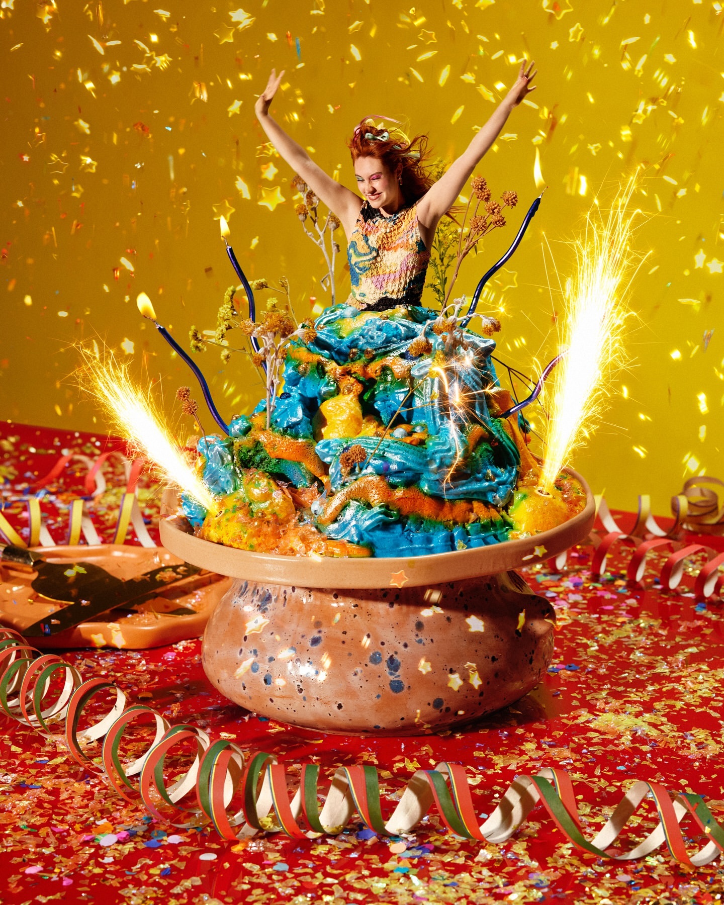 A vibrant image features a woman standing behind a colorful cake with sparklers, confetti, and streamers, set against a yellow background.