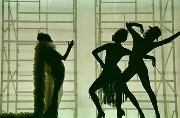 Three dancers in silhouette with geometric background. One adult female in a boa smoking, with two other dancers in motion.