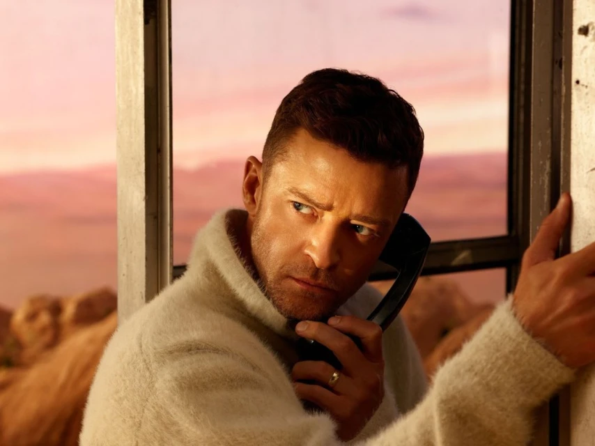 Close-up shot of an adult male in a white sweater using a black telephone inside of a tan-colored phone booth against a dusk sky.