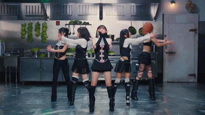 Five young adult women perform a dance routine in a commercial kitchen, wearing black and white outfits with black knee-high boots, eye-level shot.