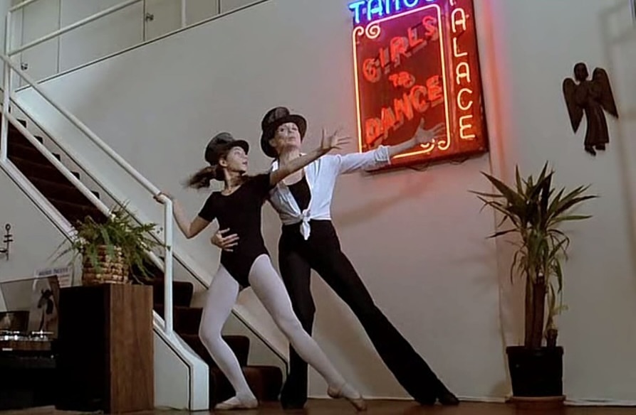 Two women dancing in a studio, one wearing a black leotard and one in a white shirt and black pants, in front of a neon sign.