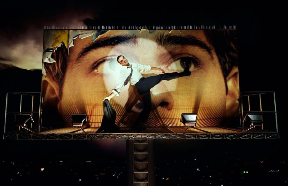 Man balances on a billboard at night, wearing a shirt and slacks. The billboard features a close-up of a man's eye.