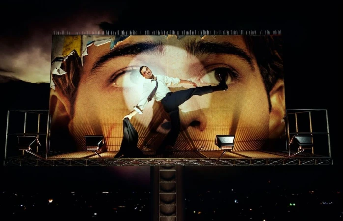 Man balances on a billboard at night, wearing a shirt and slacks. The billboard features a close-up of a man's eye.