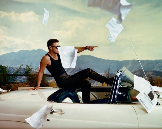 Man in a black tank top and sunglasses seated on the hood of a white convertible car as papers flutter through the air against a mountain backdrop.
