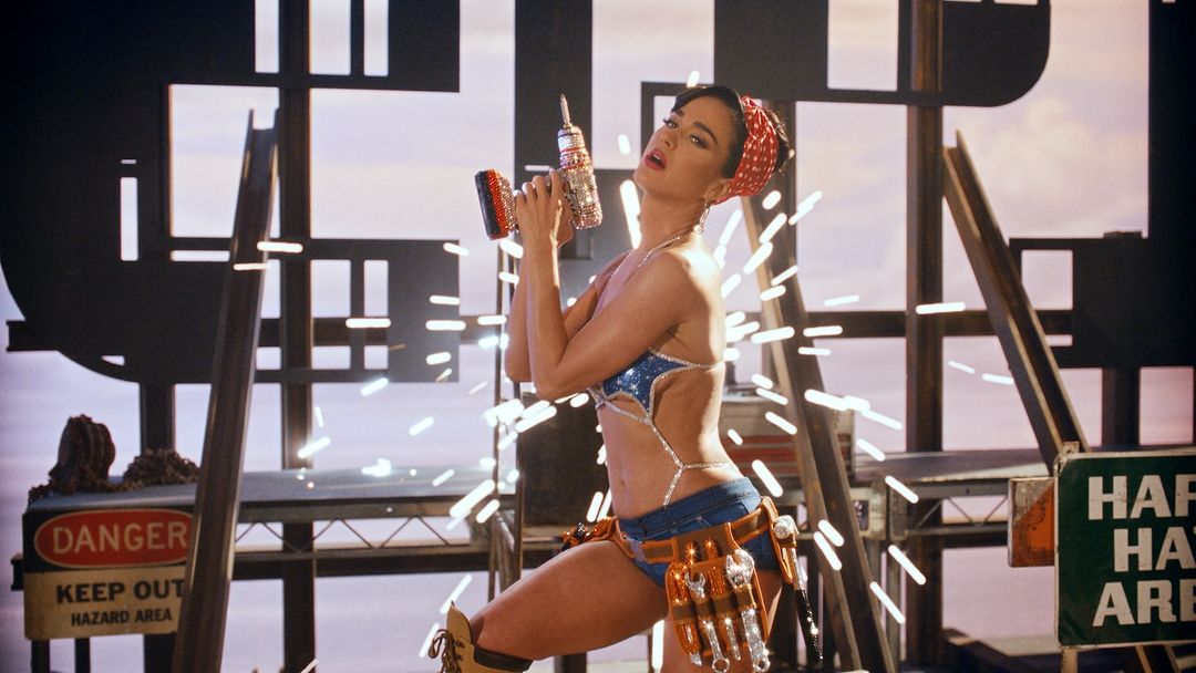 Woman poses in a bedazzled bikini and tool belt, holding a drill against a construction-themed set with warning signs and metal scaffolding.