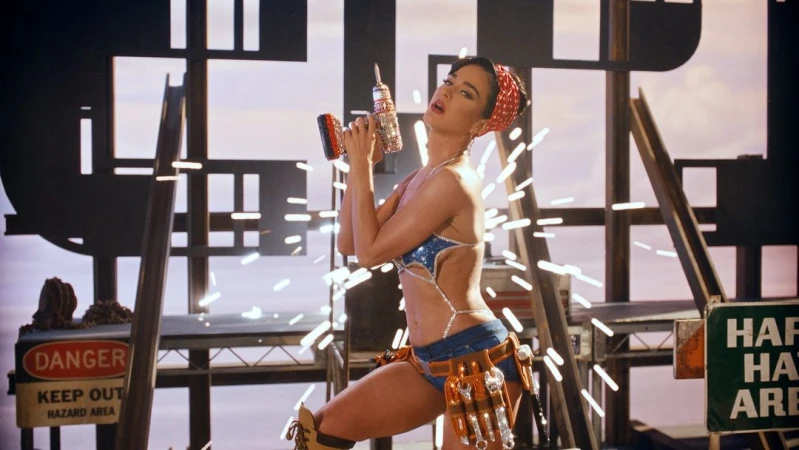 Woman poses in a bedazzled bikini and tool belt, holding a drill against a construction-themed set with warning signs and metal scaffolding.
