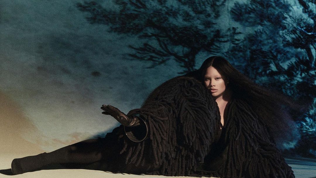 Adult woman with long black hair poses wearing a heavy textured black coat in a studio against a painted blue forest backdrop.
