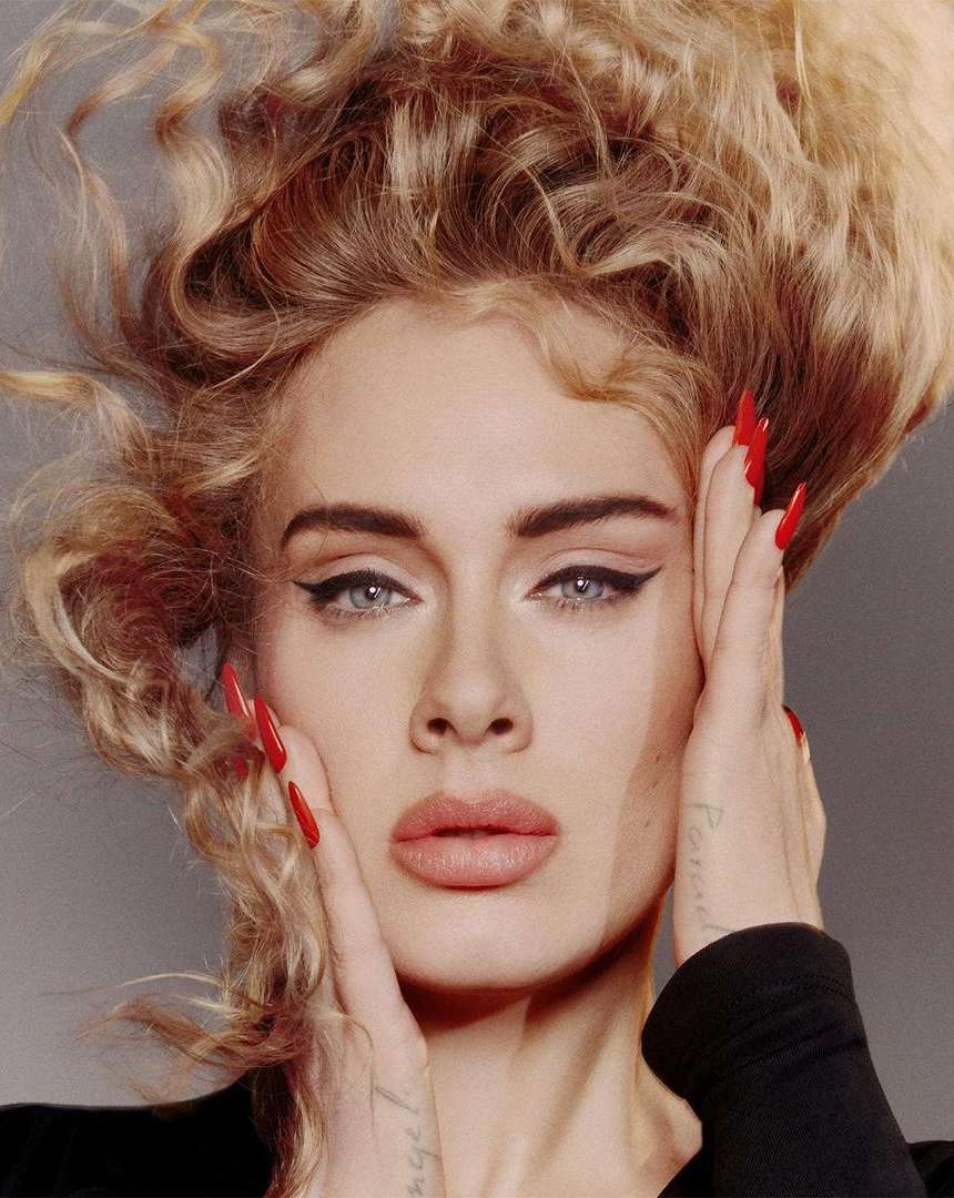 Close-up portrait of Adele with voluminous blonde hair, winged eyeliner, and long red nails framing her face by Charlotte Wales.
