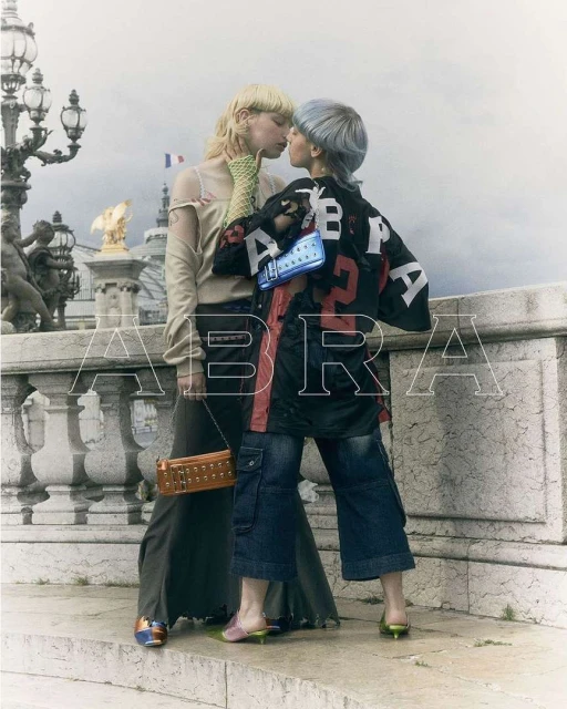 Two people kiss on a bridge in Paris, France. They wear unique street style fashion with a French flag and architecture in the background.
