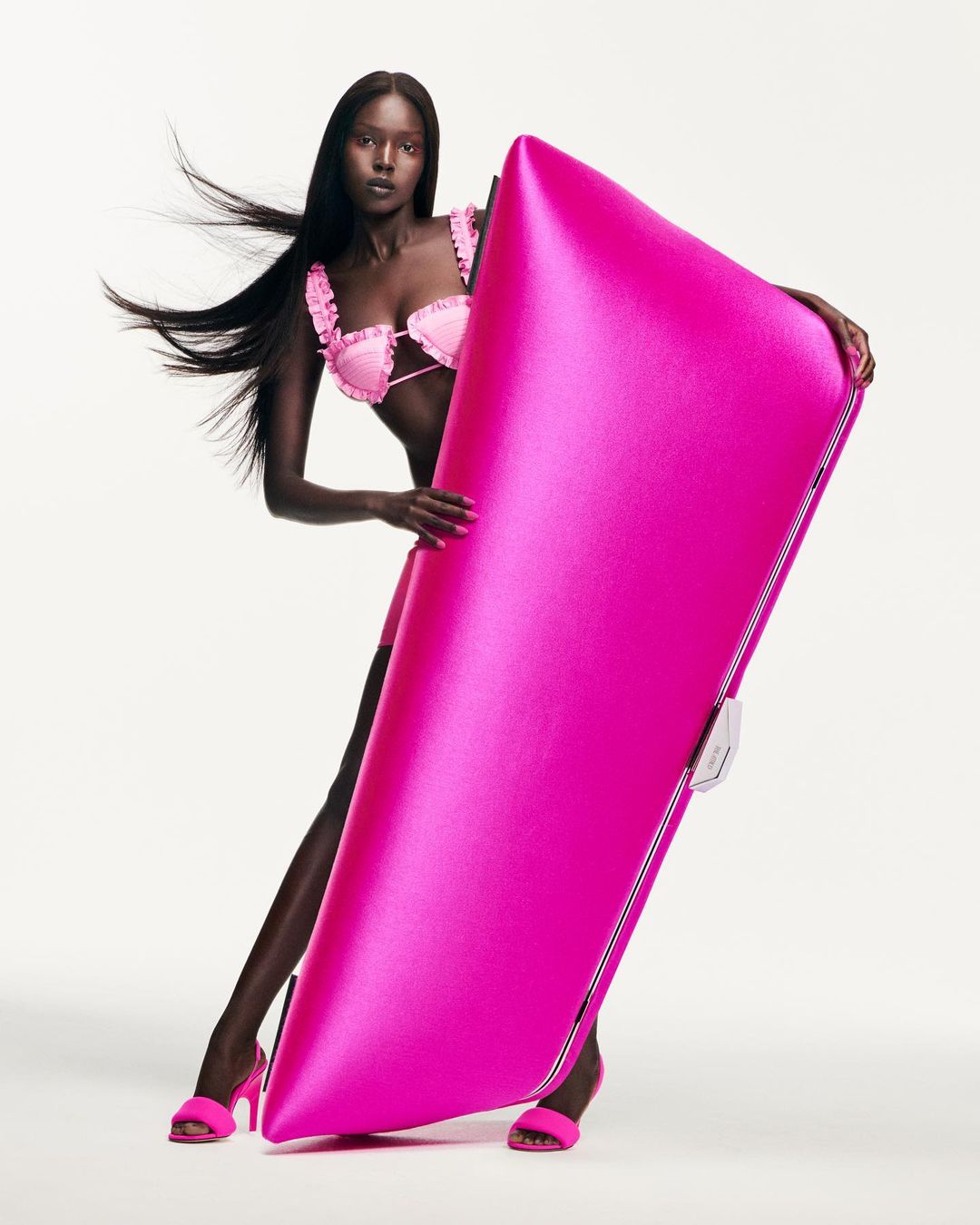 Fashion shot of a Black female model in a pink lingerie set posing with a shiny pink material against a clean white backdrop, studio lighting.