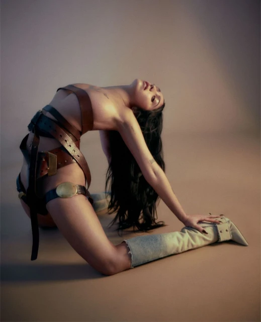 A woman in an arched pose wears leather harnesses and high boots in a studio setting with warm lighting and neutral backdrop.