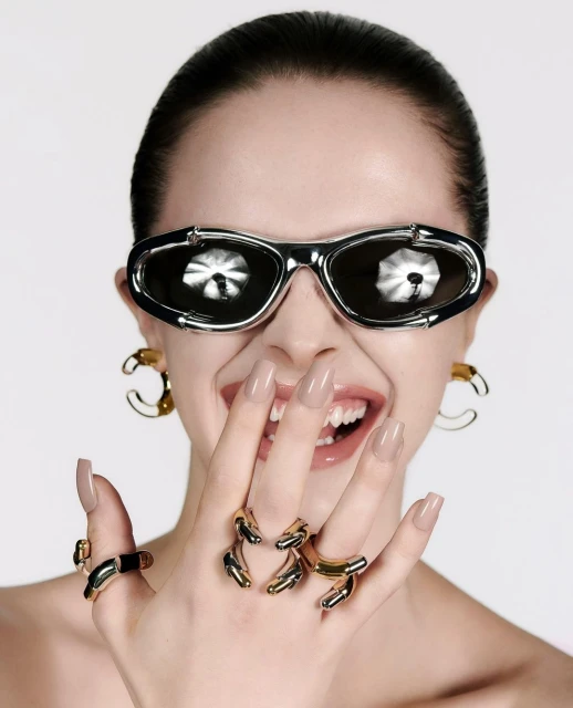 Young woman in futuristic sunglasses and gold jewelry smiles in a studio setting, showcasing a beauty and fashion aesthetic.