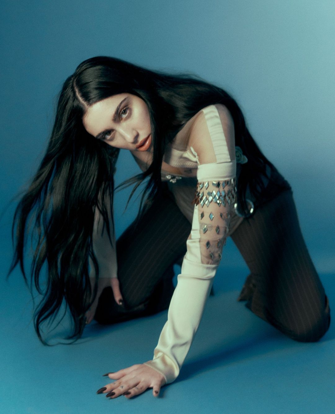 Brunette woman in tan and sheer clothing poses on her hands and knees against a gradient light-blue backdrop in a studio setting.