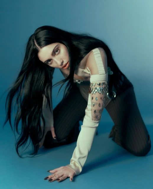 Brunette woman in tan and sheer clothing poses on her hands and knees against a gradient light-blue backdrop in a studio setting.